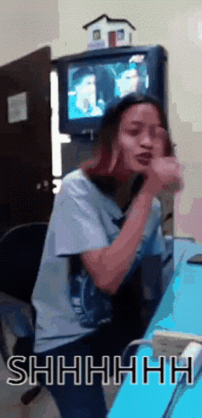 a woman is sitting at a desk in front of a television and making a shhh gesture
