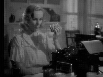A Woman Is Sitting At A Desk In Front Of A Typewriter And Holding Something In Her Hand GIF