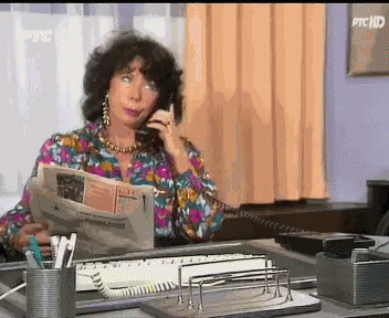 A Woman Is Sitting At A Desk Talking On A Phone And Reading A Newspaper GIF