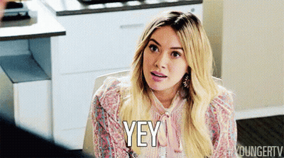 A Woman Is Sitting At A Table And Saying Yey GIF