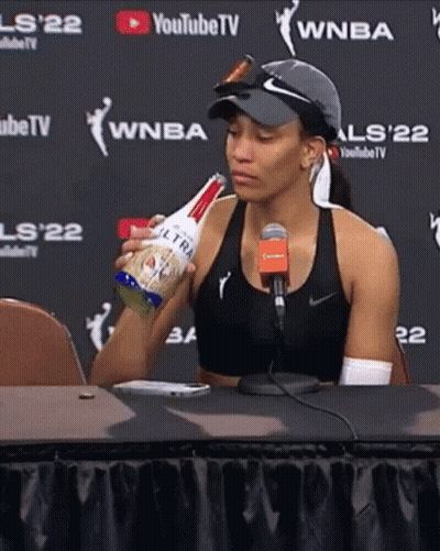 A Woman Is Sitting At A Table Holding A Bottle Of Ultra Light Beer GIF