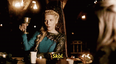 A Woman Is Sitting At A Table Holding A Goblet And The Word Skol Is Next To Her GIF