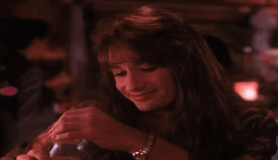 A Woman Is Sitting At A Table In A Dark Room Holding A Glass And Smiling . GIF