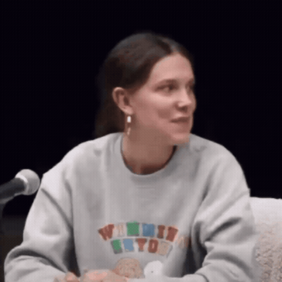 A Woman Is Sitting At A Table In Front Of A Microphone Wearing A Sweatshirt And Earrings . GIF