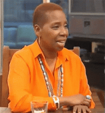 A Woman Is Sitting At A Table Wearing An Orange Shirt And Smiling . GIF