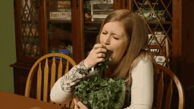 A Woman Is Sitting At A Table With A Bowl Of Greens In Front Of A Bookcase With Twenty-One On It GIF