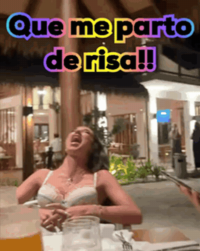 A Woman Is Sitting At A Table With A Glass Of Beer And A Sign That Says Que Me Parto De Risa !! GIF
