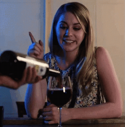 A Woman Is Sitting At A Table With A Glass Of Wine And A Bottle Of Wine Being Poured Into It GIF