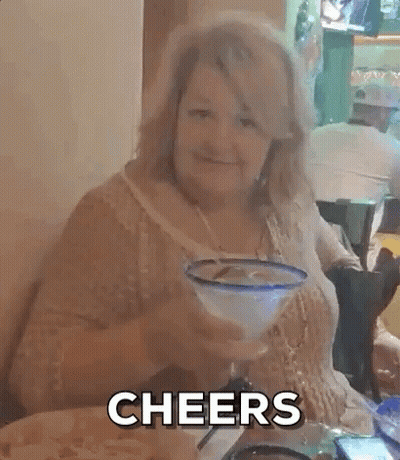 A Woman Is Sitting At A Table With A Margarita In Her Hand And The Words Cheers Behind Her . GIF