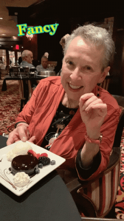 A Woman Is Sitting At A Table With A Plate Of Dessert In Front Of A Sign That Says Fancy GIF