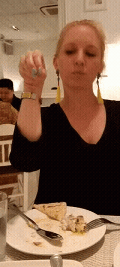 A Woman Is Sitting At A Table With A Plate Of Food And A Fork In Her Hand GIF