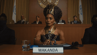 A Woman Is Sitting At A Table With A Sign That Says Wakanda GIF