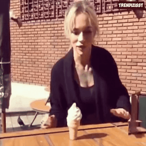 A Woman Is Sitting At A Table With An Ice Cream Cone In Front Of Her And The Words Trendizisst On The Bottom GIF