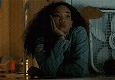 A Woman Is Sitting At A Table With Her Hand On Her Face And A Fan In The Background . GIF