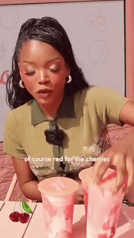 A Woman Is Sitting At A Table With Two Cups Of Cherry Juice GIF