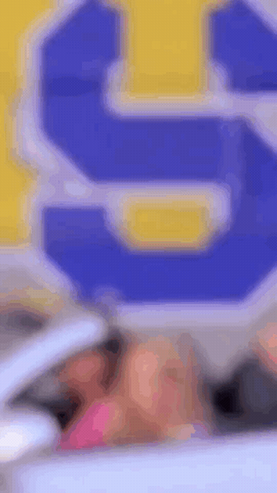 A Woman Is Sitting In A Car In Front Of A Blue And Yellow Sign That Says S. GIF