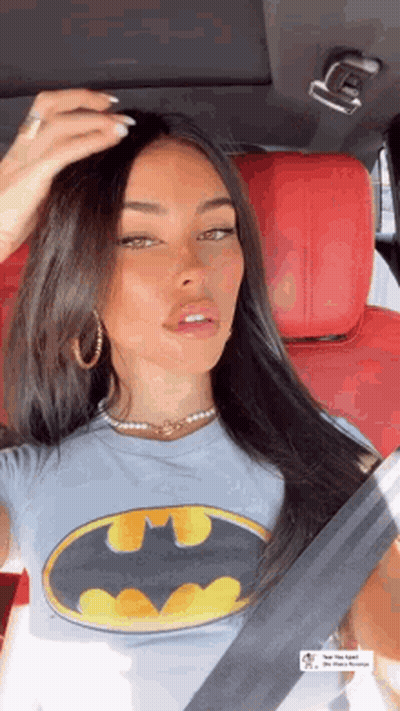 A Woman Is Sitting In A Car Wearing A Batman T-Shirt And Earrings . GIF