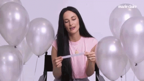A Woman Is Sitting In A Chair Surrounded By Silver Balloons And Holding A Piece Of Paper . GIF