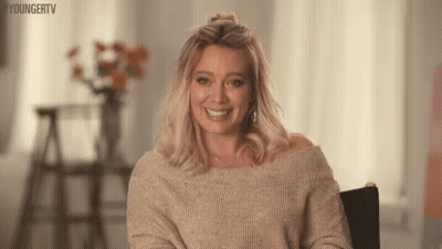 A Woman Is Sitting In A Chair Wearing A Sweater And Smiling . GIF