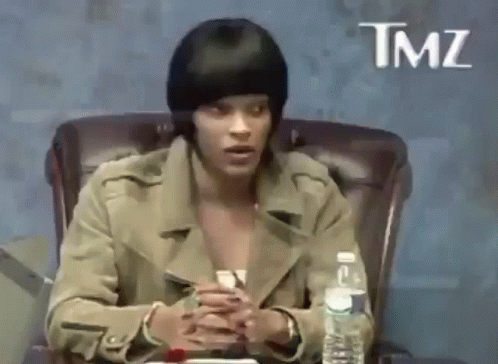 A Woman Is Sitting In A Chair With A Bottle Of Water In Front Of Her And A Sign That Says Tmz . GIF