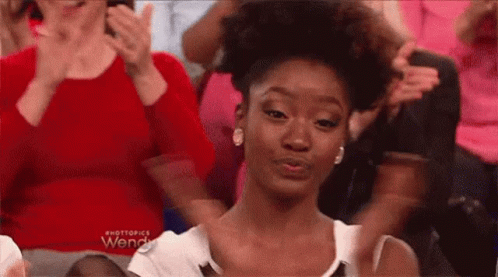A Woman Is Sitting In A Crowd Of People Clapping And Making A Funny Face . GIF