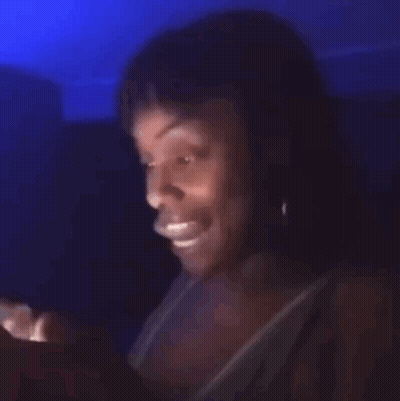 A Woman Is Sitting In A Dark Room Looking At A Cell Phone . GIF