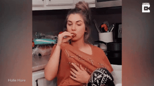 A Woman Is Sitting In A Kitchen Eating A Piece Of Food And The Words Hollie Haine Are On The Bottom GIF