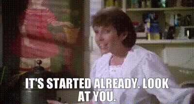 A Woman Is Sitting In A Kitchen With The Words `` It 'S Started Already Look At You '' Written On The Screen . GIF