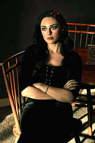 A Woman Is Sitting In A Wooden Chair With Her Arms Crossed In Front Of A No Smoking Sign GIF