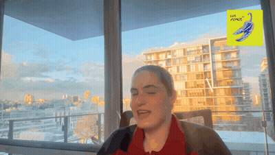 A Woman Is Sitting In Front Of A Window With A Yellow Sign That Says Love Plumes On It GIF