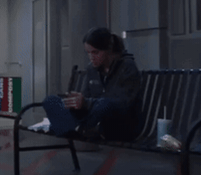 A Woman Is Sitting On A Bench Looking At Her Cell Phone GIF