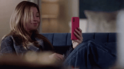 A Woman Is Sitting On A Blue Couch Looking At Her Phone GIF