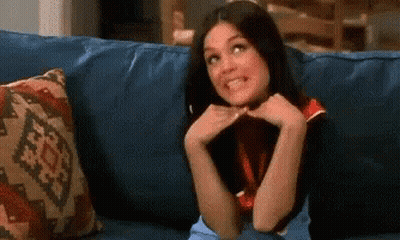 A Woman Is Sitting On A Blue Couch With Her Hands On Her Face And Smiling . GIF