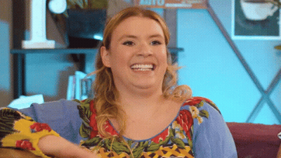 A Woman Is Sitting On A Couch And Smiling While Wearing A Colorful Shirt GIF