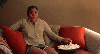 A Woman Is Sitting On A Couch Eating Popcorn And Making A Surprised Face . GIF