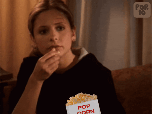 A Woman Is Sitting On A Couch Eating Popcorn From A Container That Says Pop Corn GIF