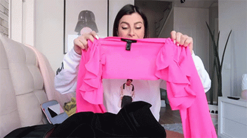 A Woman Is Sitting On A Couch Holding A Bright Pink Ruffled Top . GIF