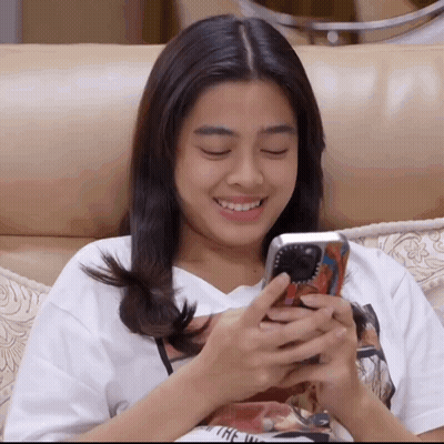 A Woman Is Sitting On A Couch Looking At Her Phone And Smiling GIF