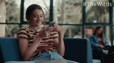 A Woman Is Sitting On A Couch Looking At Her Phone With #Thewilds Written In The Corner GIF