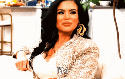 A Woman Is Sitting On A Couch Wearing A Sequined Jacket And Earrings And Saying `` I 'M '' . GIF