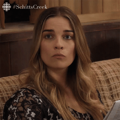 A Woman Is Sitting On A Couch With A Newspaper In Her Hand And The Word Schitts Creek On The Bottom GIF