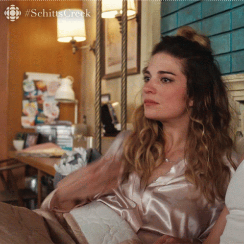 A Woman Is Sitting On A Couch With A #Schitts Creek Logo On The Bottom GIF