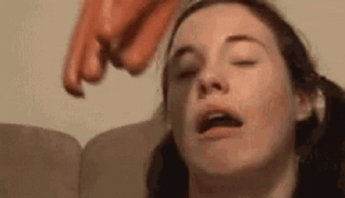 A Woman Is Sitting On A Couch With Her Eyes Closed While A Hot Dog Is Being Thrown At Her Head . GIF