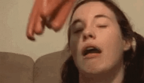 A Woman Is Sitting On A Couch With Her Eyes Closed While A Sausage Is Being Thrown At Her Head . GIF