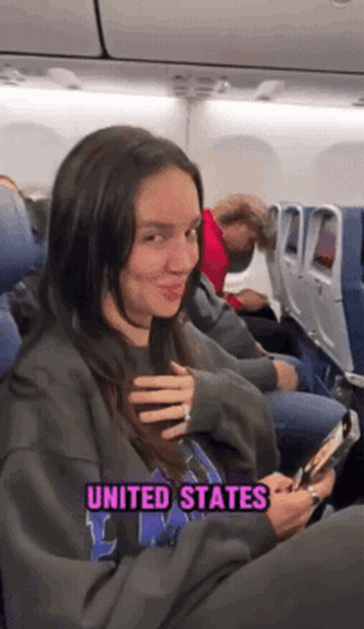 A Woman Is Sitting On An Airplane Wearing A Sweatshirt That Says United States On It . GIF