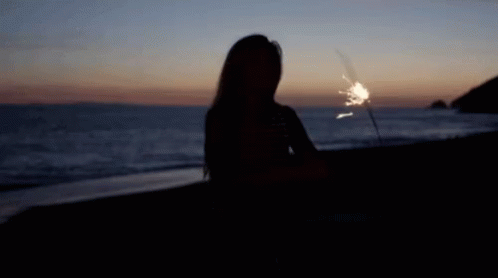A Woman Is Sitting On The Beach Holding A Sparkler At Sunset . GIF