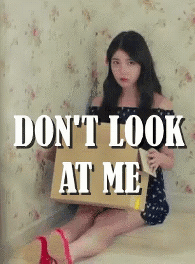 A Woman Is Sitting On The Floor Holding A Cardboard Box That Says " Don T Look At Me " GIF
