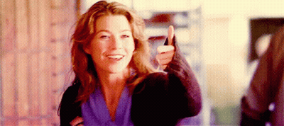 A Woman Is Smiling And Giving A Thumbs Up Sign While Standing In Front Of A Brick Wall . GIF