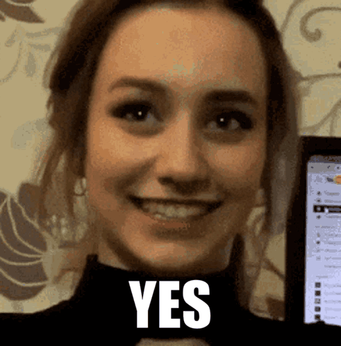 A Woman Is Smiling And Holding A Cell Phone In Front Of A Screen That Says Yes . GIF