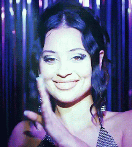 A Woman Is Smiling And Waving Her Hand In Front Of A Curtain GIF